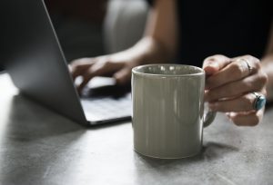 Eine Person tippt auf einen Laptop und greift mit der anderen Hand eine Tasse. 